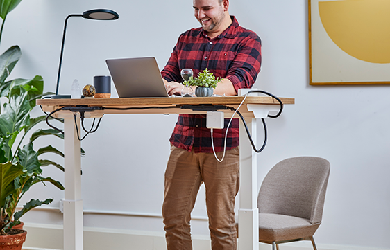 A man standing at his desk, Facetiming a friend, with a computer, light, and mug on the desk, next to the Table Top Power.